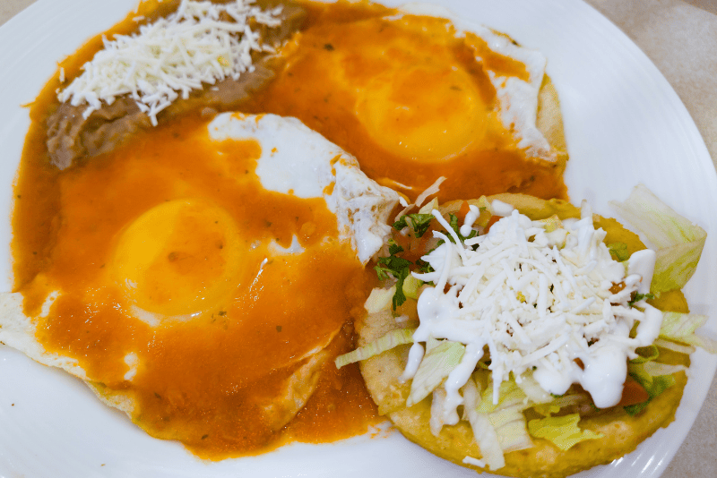 Huevos fritos sobre tortilla, cubiertos con salsa roja espesa y acompañados de frijoles refritos.