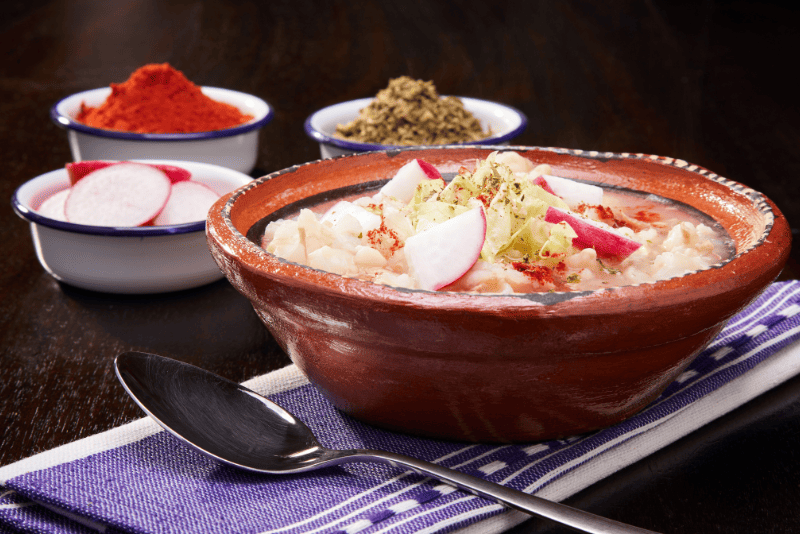 Tazón humeante de pozole rojo con maíz cacahuazintle, carne y guarniciones frescas.